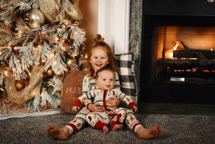 Brother and sister, little boy and girl posing in matching pajamas in front of christmas tree and fireplace