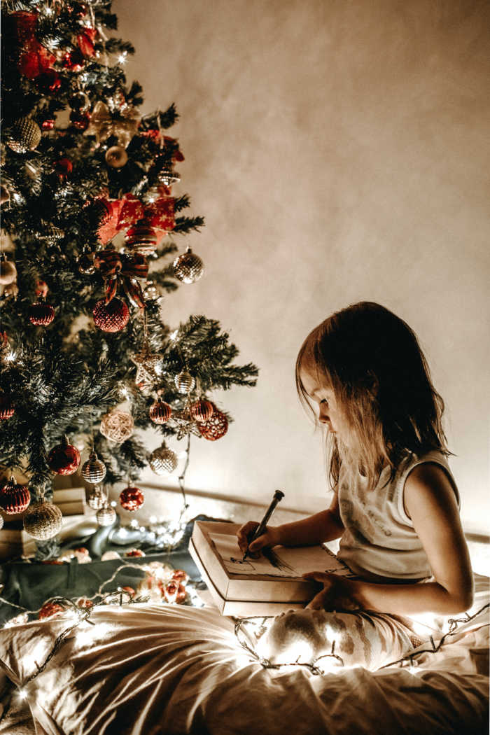 little girl lit with christmas lights by christmas tree writing to Santa