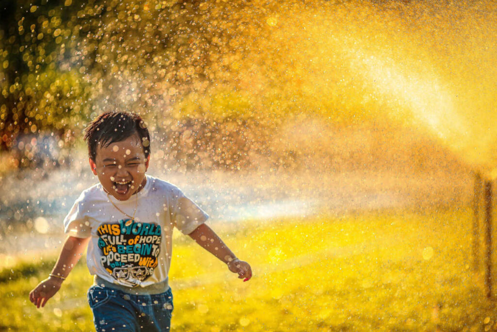 boy laughing running through sprinkler in his clothes