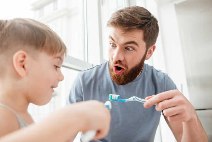 Father and son smiling while brushing teeth in bathroom. Look at each other.