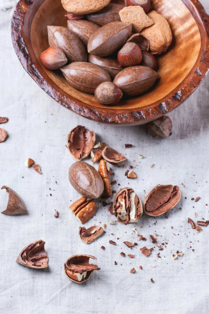 Wooden bowl with whole and chopped pecan nuts over gray textile.
